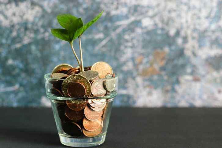 A small green plant sprout growing out of a glass jar filled with various gold and copper coins, set against a dark surface and a textured blue-grey background.
